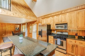 A kitchen with wooden cabinets, a granite countertop, a refrigerator, a microwave, a stove, and a dining area with chairs.