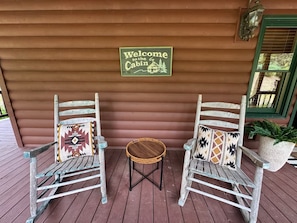 Two rustic wooden rocking chairs with patterned cushions on a wooden deck, a welcome sign, and a green-framed window.