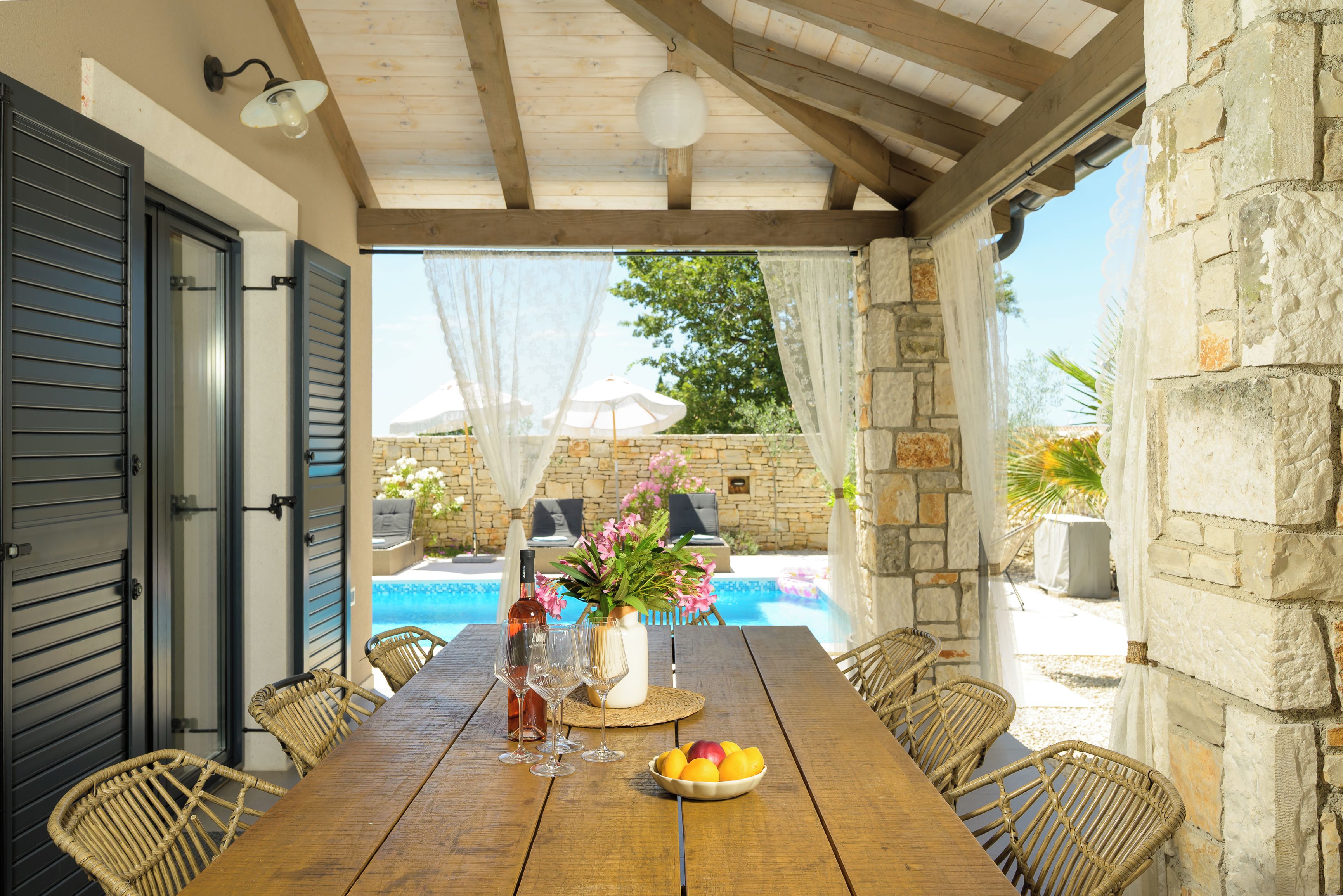 An outdoor dining area with a wooden table, wicker chairs, a stone wall, and a view of a swimming pool.