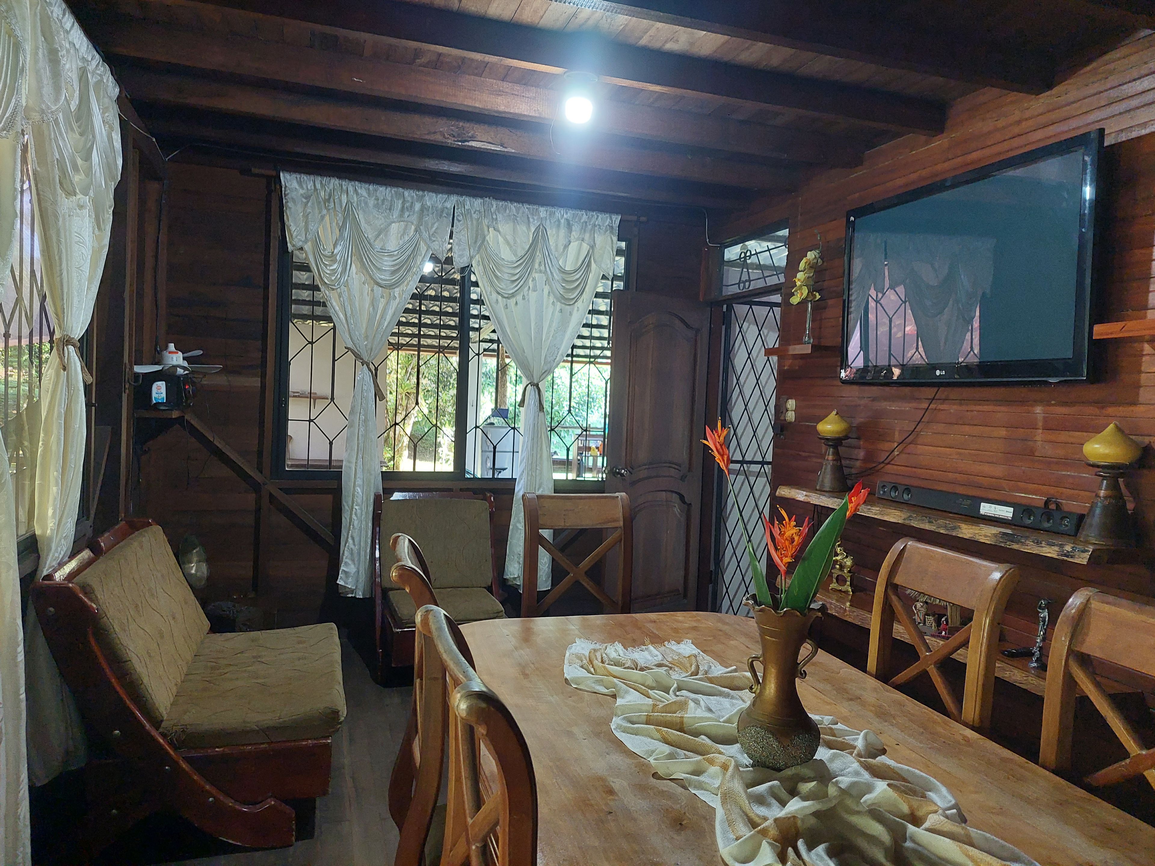 A wooden interior with a dining table, chairs, a TV mounted on the wall, and a window with curtains.