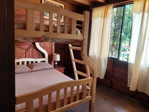A wooden bunk bed with a ladder, a bedside table with a lamp, and a window with curtains.
