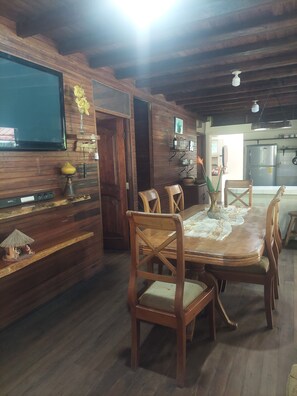 Un comedor de madera con una mesa y sillas, un televisor montado en la pared y una cocina al fondo.