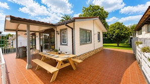 A covered outdoor dining area with a wooden table and benches, a pergola, and a view of a residential building.