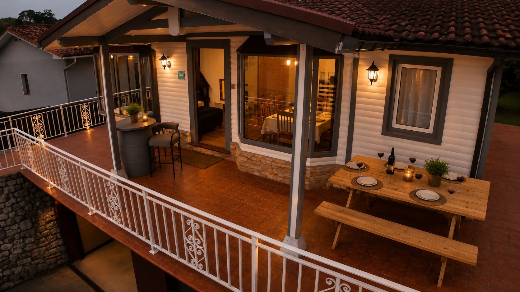 A covered outdoor dining area with a wooden table and benches, a stone wall, and a balcony railing.