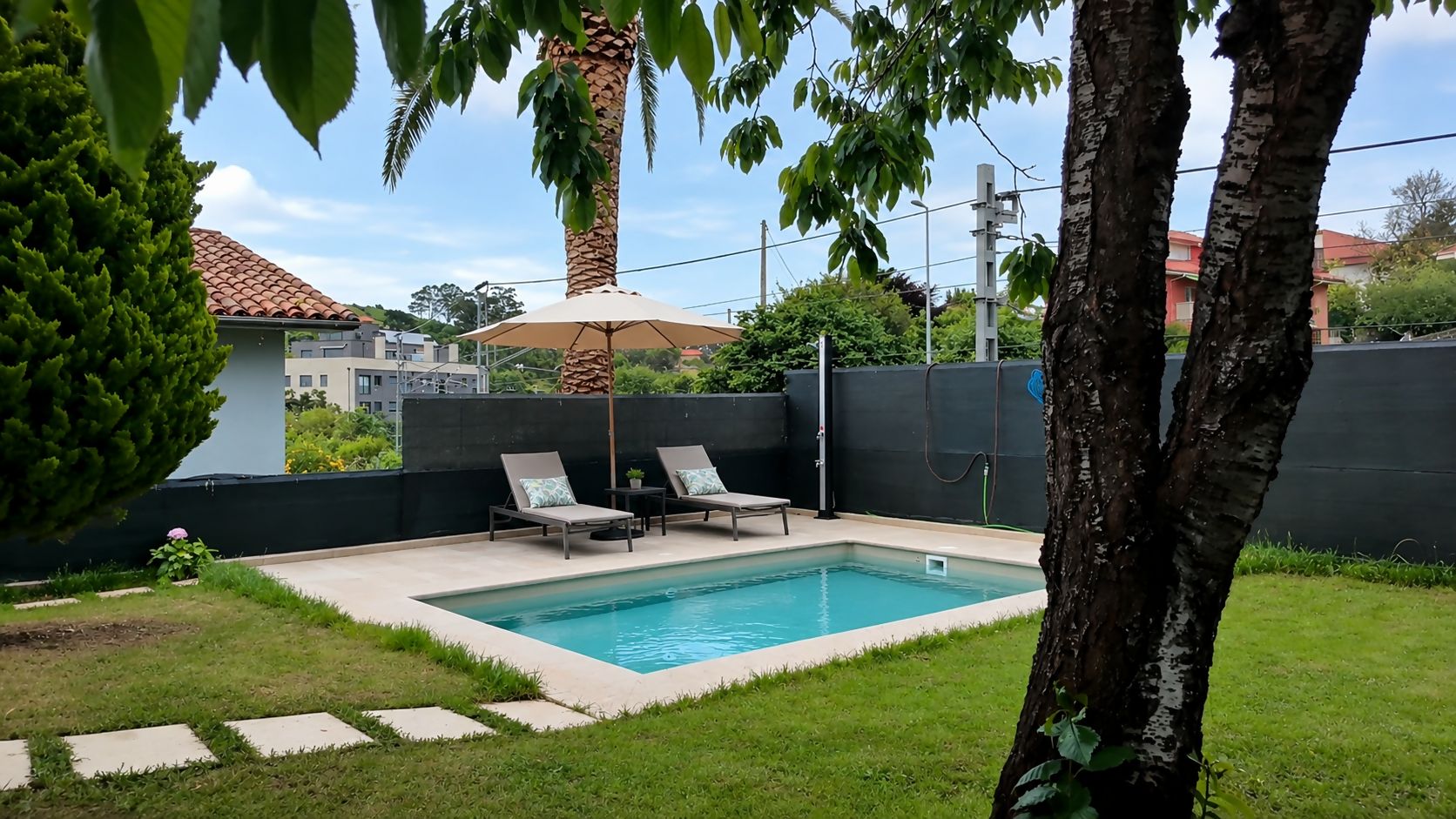 Une piscine rectangulaire, agrémentée de deux chaises longues et d’une table, le tout entouré d’un petit muret et de verdure.