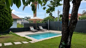 A rectangular swimming pool with two lounge chairs and a table, surrounded by a low wall and greenery.