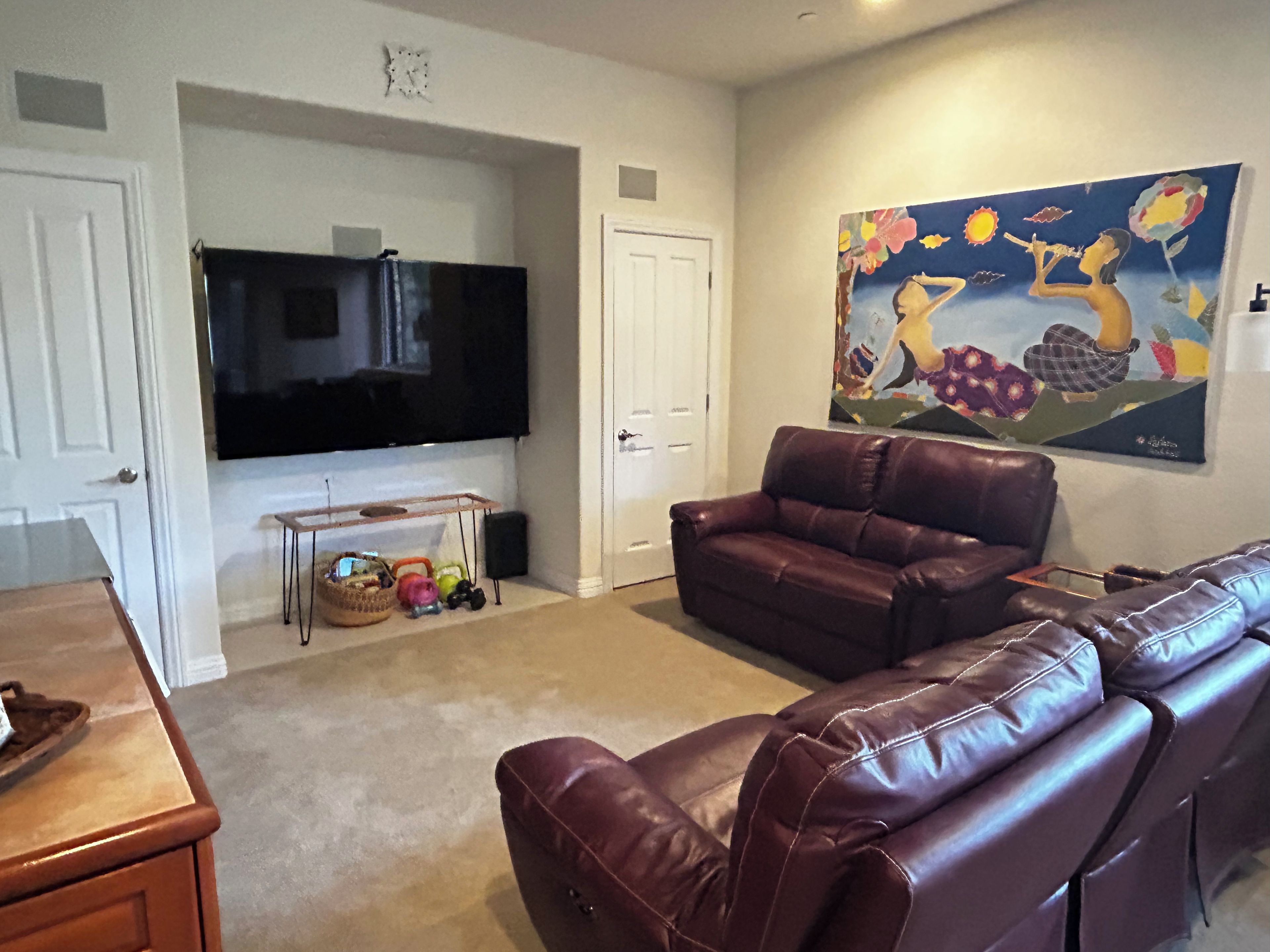 A living room with a large flat-screen TV, a brown leather sofa, a coffee table, and a painting on the wall.