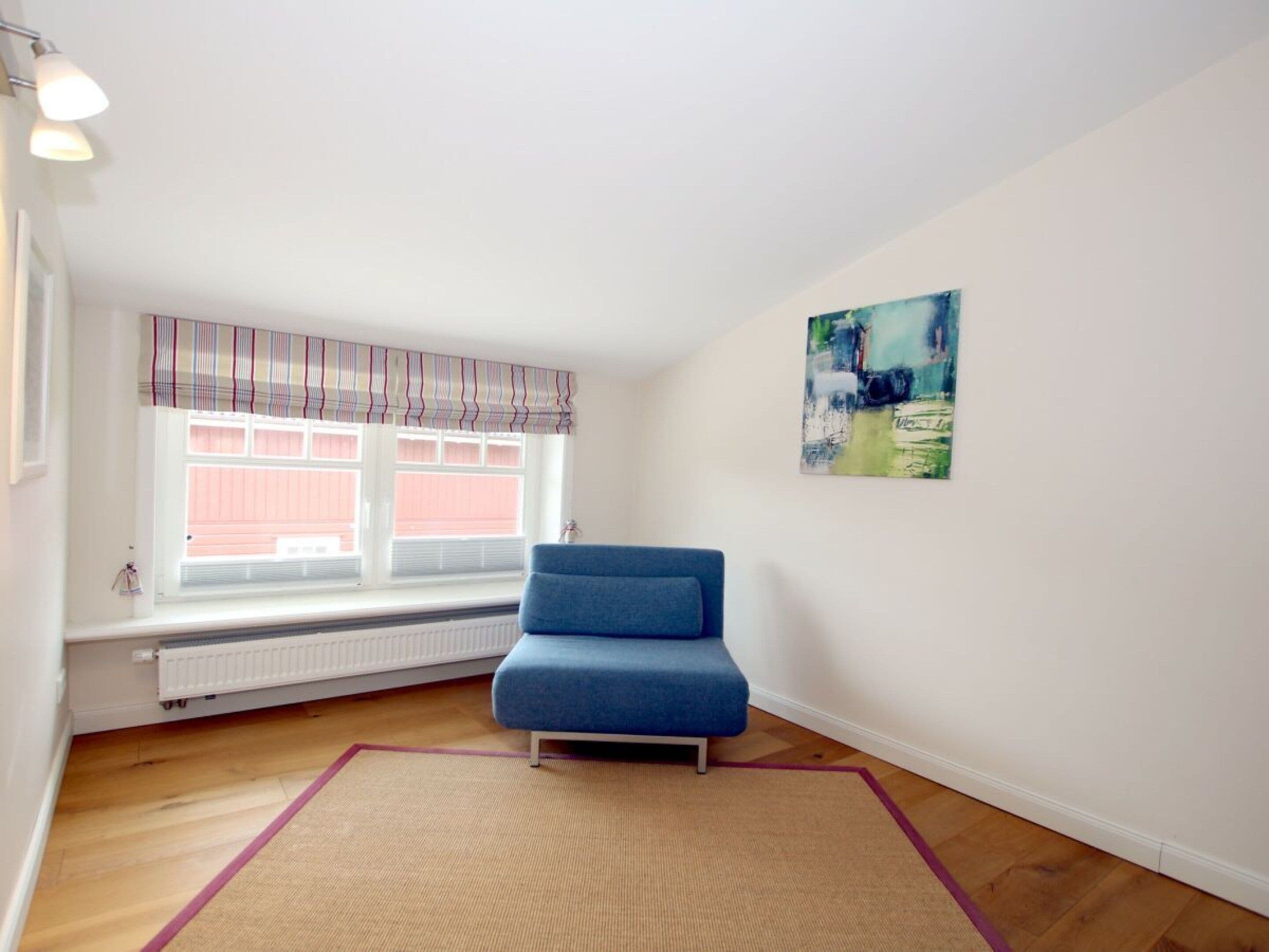 Une pièce avec un canapé bleu, un radiateur, une fenêtre avec des stores et un tableau accroché au mur.