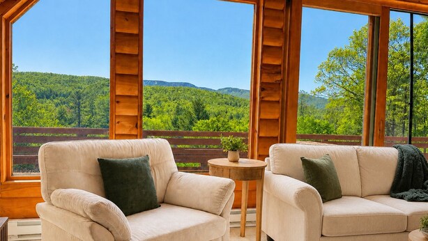 A room with two beige armchairs, a small round table with a plant, and a view of a forested landscape through large windows. - Luxury Lodge for 2–4 • Gore Views , AC, Pets (North Creek)
