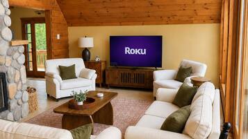 Een gezellige woonkamer met een stenen openhaard, witte banken, een houten salontafel en een televisie waarop 'Roku' wordt weergegeven.