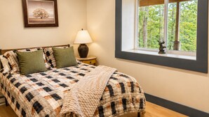 A bedroom with a bed, a nightstand, a lamp, a window with a view of trees, and a framed picture on the wall. - Luxury Lodge for 2–4 • Gore Views , AC, Pets (North Creek)