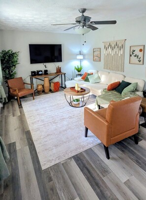 A living room with a ceiling fan, a flat-screen TV, a sofa, a coffee table, and a patterned rug. - Delightful 2-bedroom house with an office, deck and fire pit in Sioux Falls (Sioux Falls)