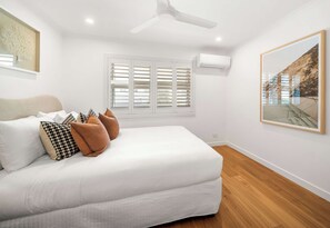 A bedroom with a bed, white walls, a ceiling fan, a window with shutters, and a framed picture on the wall.