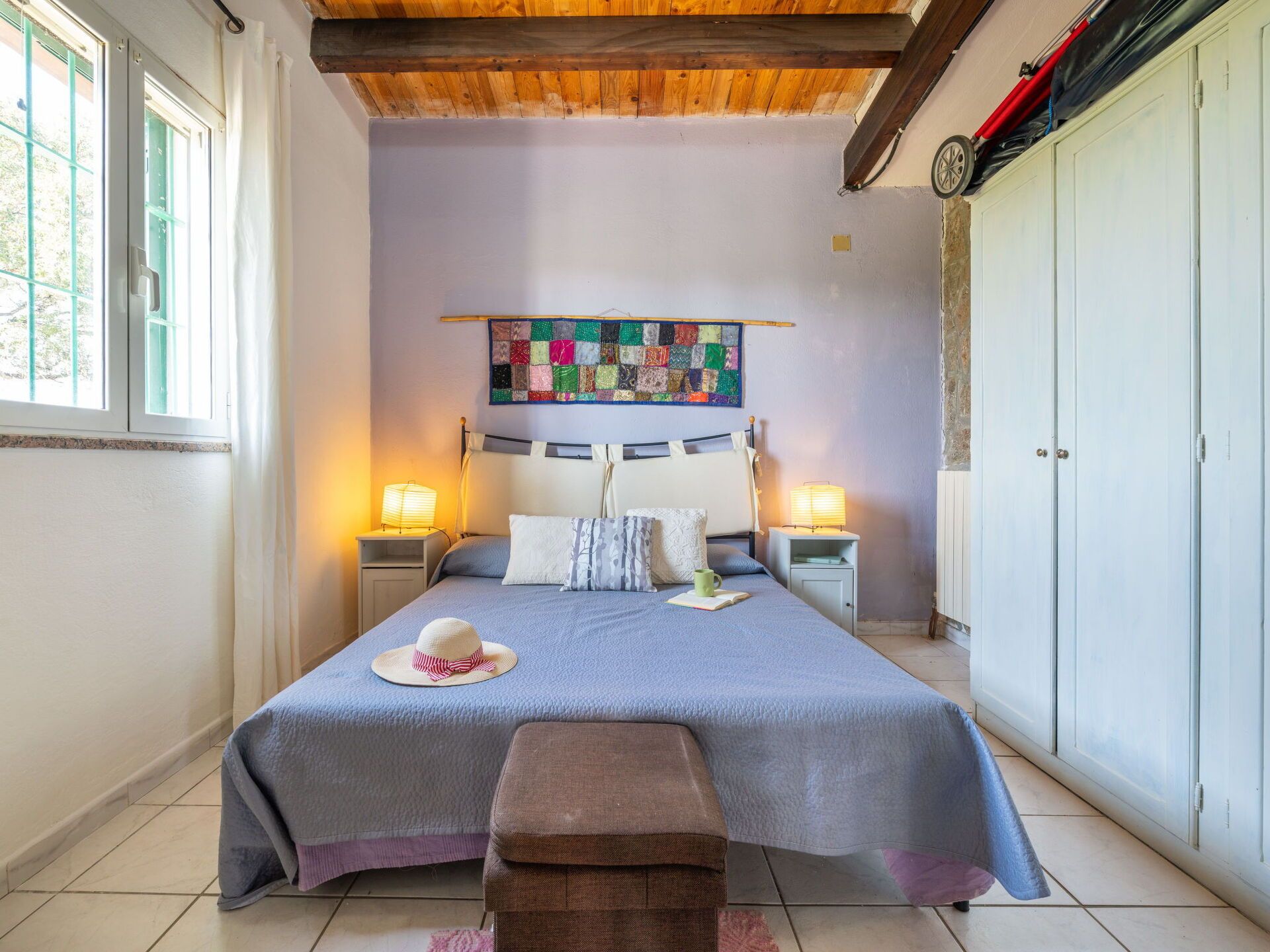 A bedroom with a bed, two bedside tables, a bench, a quilted headboard, a wall hanging, and a wardrobe.