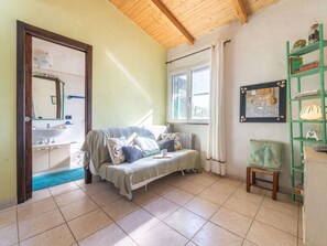 Ein gemütliches Zimmer mit Bett, kleinem Sessel, einer Bücherablage, einem Fenster und einem Badezimmer mit Waschbecken und Spiegel.