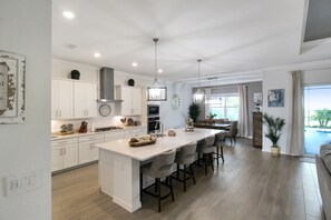 A modern kitchen with white cabinetry, a central island, and a dining area with a view of the outdoors. - Pool Home In Calusa Golf & Country Club - Fore Seasons (Bradenton)