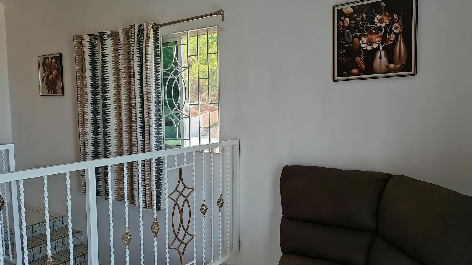 Une pièce avec un canapé marron, une rambarde blanche, une fenêtre avec des rideaux à motifs et un tableau encadré au mur.