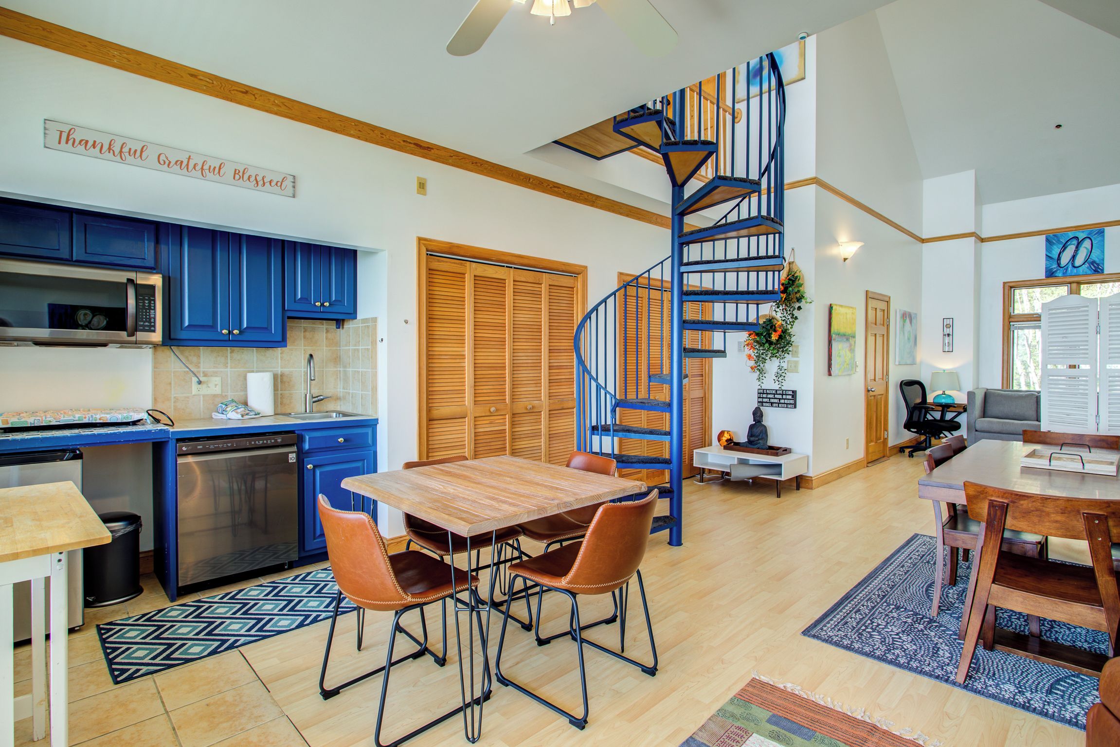 A modern kitchen with blue cabinets, a wooden dining table with chairs, and a spiral staircase.