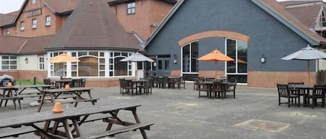An outdoor patio area with picnic tables and umbrellas, a brick building with a large overhang, and a car parked in the background.