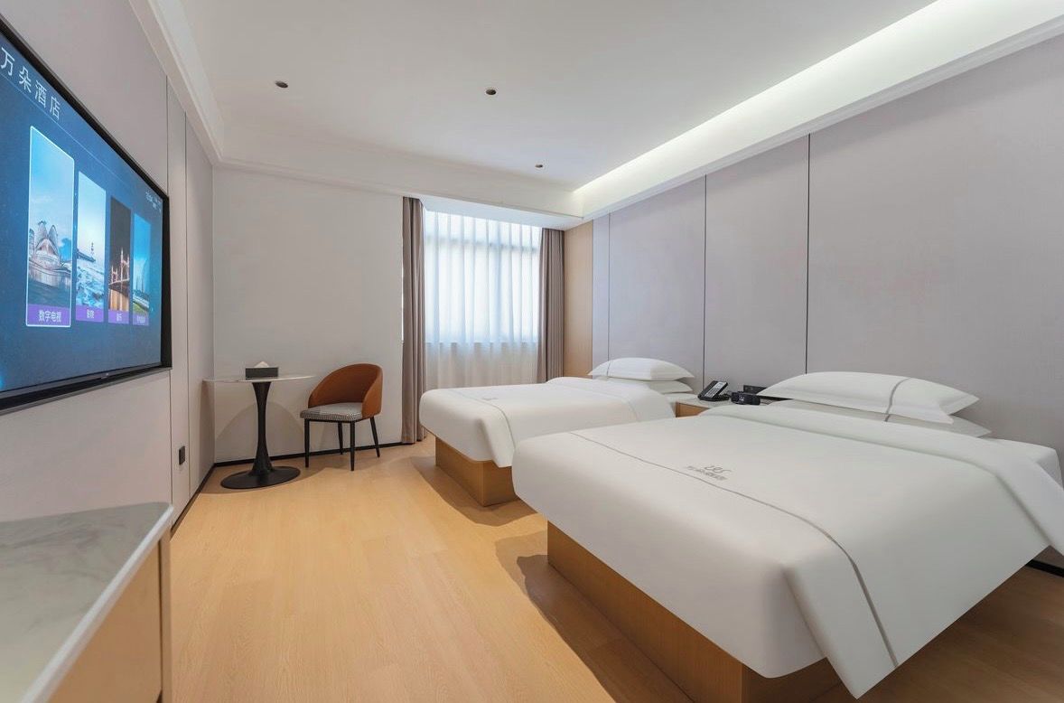 A hotel room with two beds, a desk, a chair, and a large screen displaying images.
