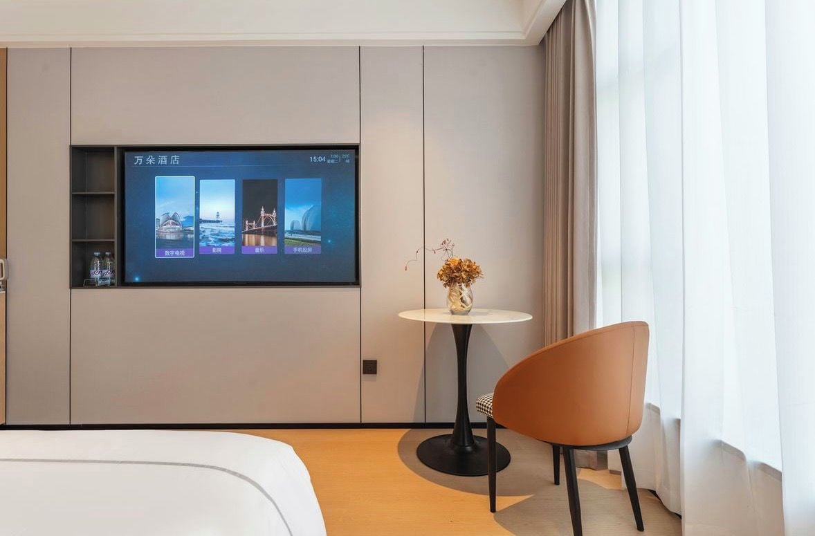 A hotel room with a bed, a small table with a vase, a TV screen displaying hotel images, and a window with curtains.