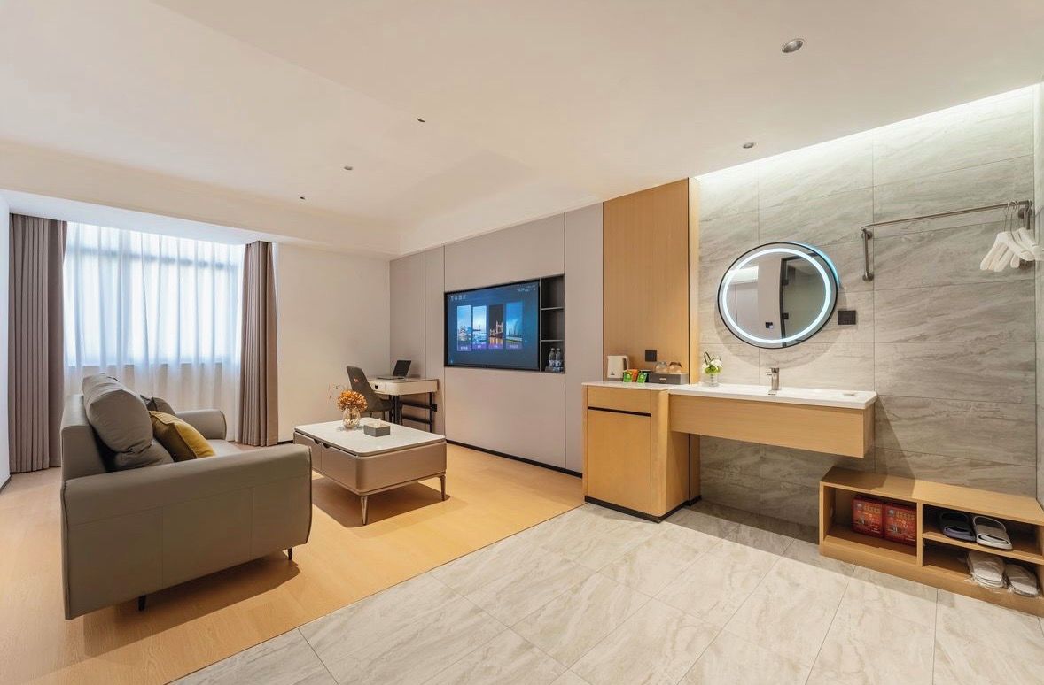 A modern hotel room with a sofa, a TV, a desk, a round mirror, and a bathroom vanity.