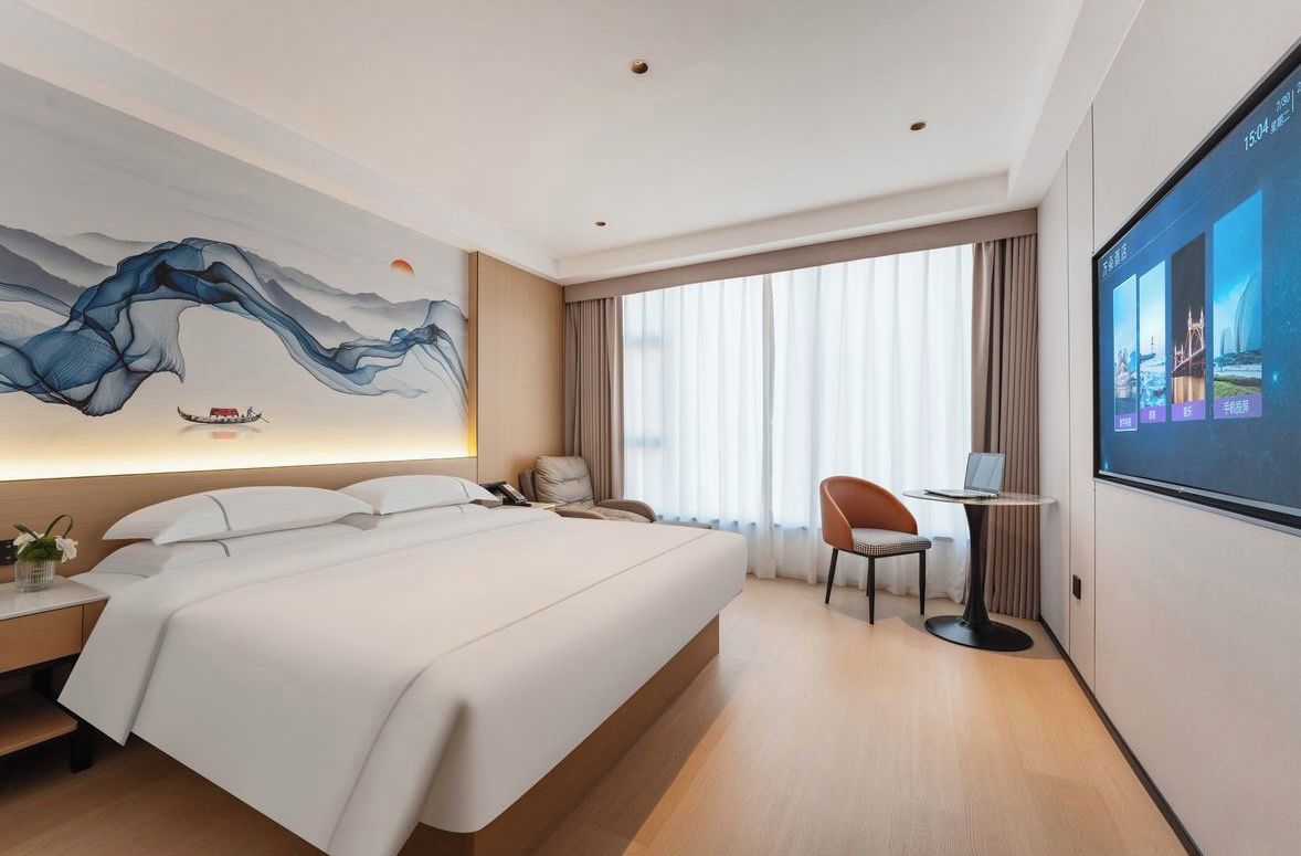 A hotel room with a large bed, a desk, a chair, and a TV screen displaying a cityscape.