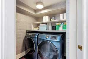 A laundry room with two front-loading washing machines and a shelf above containing folded towels and cleaning supplies. - Brick & Beam | Luxury Garden Apartment | 1855 (Savannah)