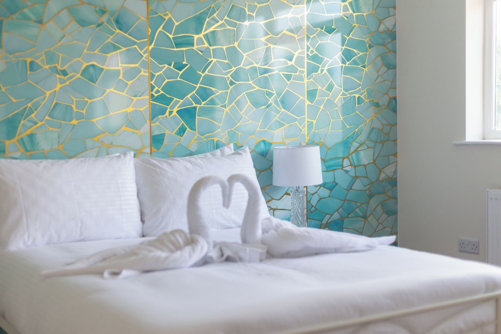 Een net opgemaakt bed met witte lakens en kussens, een bedlampje en een muur met een geometrisch patroon.