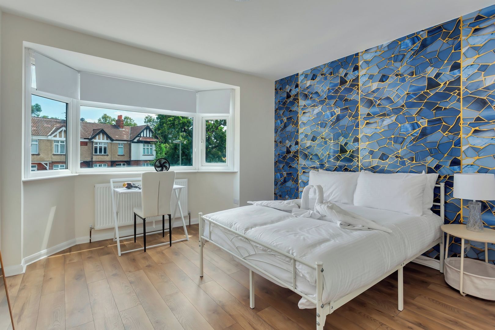 A bedroom with a bed, a desk, and a view of houses through a large window.