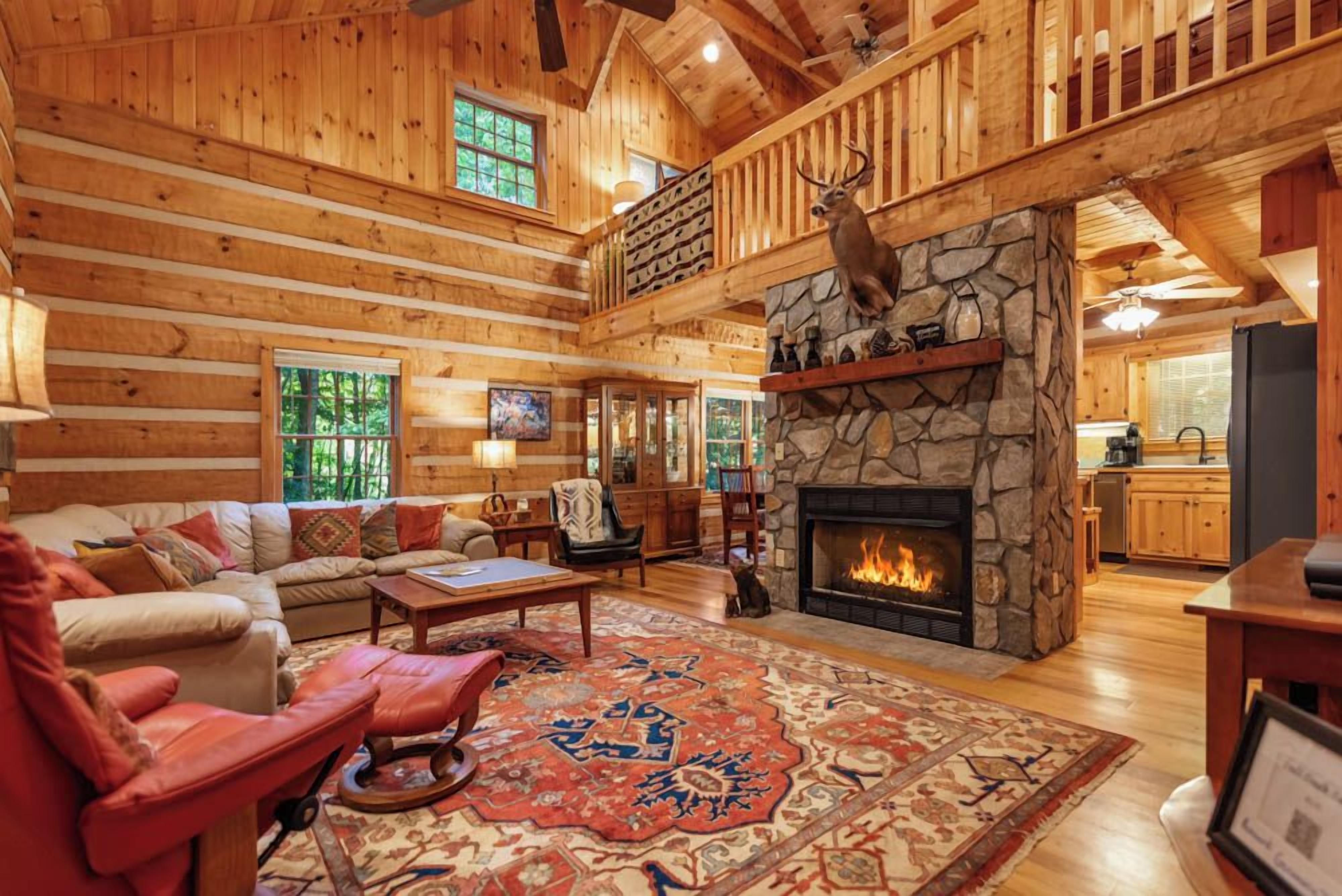 L’intérieur d’une cabane en rondins, avec une cheminée en pierre, une tête de cerf décorative accrochée au mur et un coin salon confortable comprenant un canapé et une table basse.