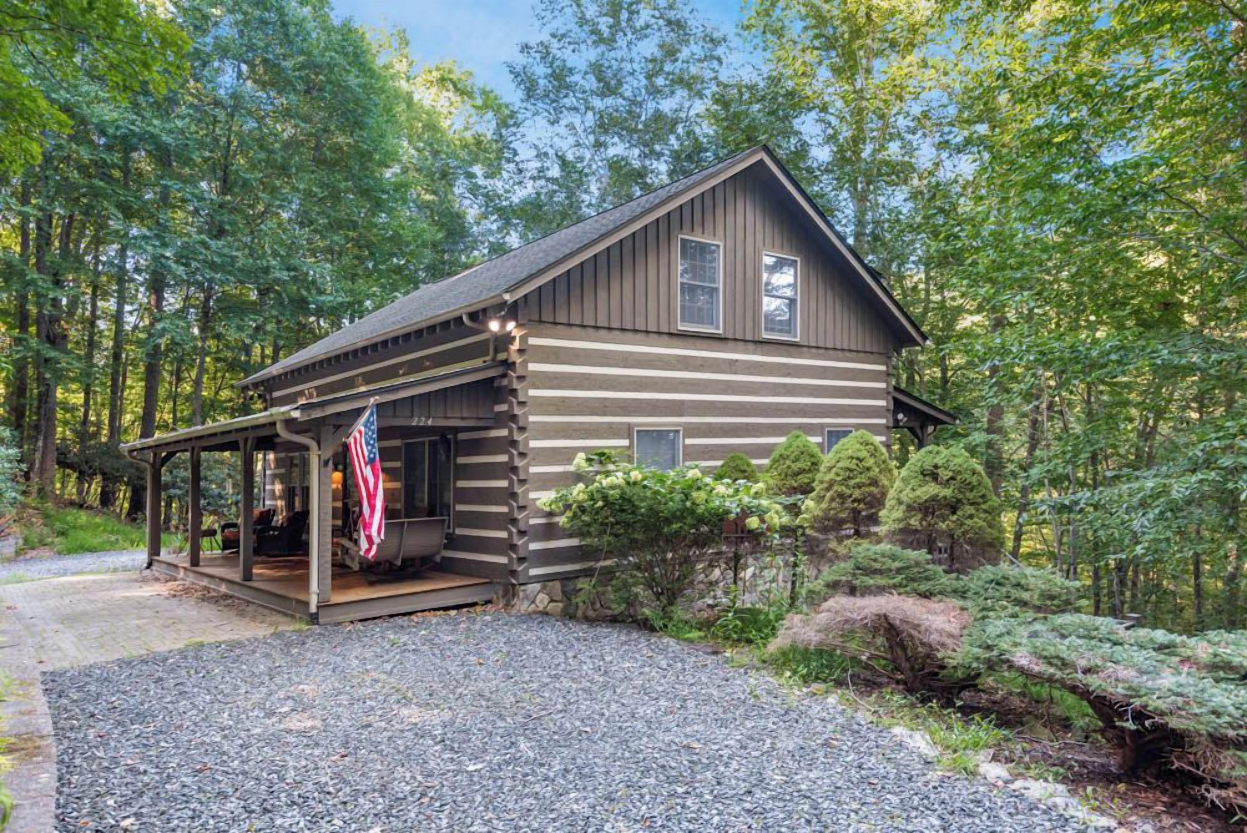 A log cabin with a porch, surrounded by trees and a gravel driveway.