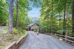 A rustic cabin with a gravel driveway, surrounded by tall trees and a wooden fence. - Todd Hideaway Overlooking Creek - Hot Tub, Fire Pit, Game Room (Todd)