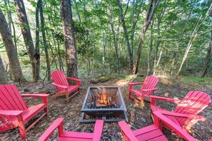 A forest setting with a fire pit and red Adirondack chairs arranged around it. - Todd Hideaway Overlooking Creek - Hot Tub, Fire Pit, Game Room (Todd)