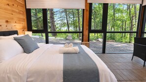 A large bed with a grey headboard, white linens, and a grey throw blanket. There are two rolled towels on the bed. The room has large windows with a view of trees and a gravel path outside. - The Lake House at Moira | 270° Lake Views, Hot Tub, Game Room & Sport Court (Centre Hastings)