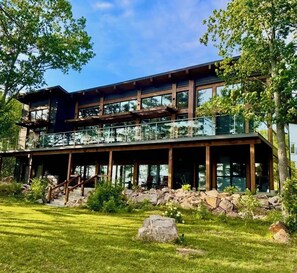 A modern two-story building with a glass balcony, surrounded by a well-maintained lawn and large rocks. - The Lake House at Moira | 270° Lake Views, Hot Tub, Game Room & Sport Court (Centre Hastings)