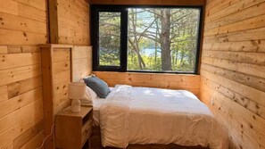 Een houten kamer met een bed, een nachtkastje en een raam met uitzicht op bomen en een wateroppervlak.