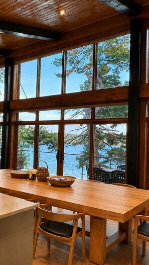 A wooden dining table with chairs, large windows, and a view of a body of water. - The Lake House at Moira | 270° Lake Views, Hot Tub, Game Room & Sport Court (Centre Hastings)