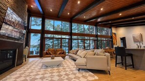 A modern living room with a fireplace, large windows, a sofa, and a kitchen area. - The Lake House at Moira | 270° Lake Views, Hot Tub, Game Room & Sport Court (Centre Hastings)