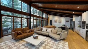 A modern living room with a sectional sofa, a coffee table, and a view of the outdoors through large windows. - The Lake House at Moira | 270° Lake Views, Hot Tub, Game Room & Sport Court (Centre Hastings)