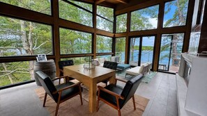 A spacious room with a large wooden dining table, black upholstered chairs, and a view of a lake through large windows. - The Lake House at Moira | 270° Lake Views, Hot Tub, Game Room & Sport Court (Centre Hastings)