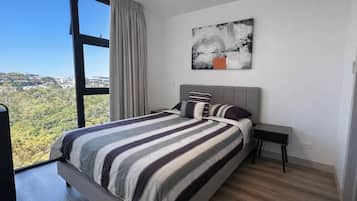 A modern bedroom with a large window, a bed with striped bedding, a nightstand, and a wall-mounted abstract painting.