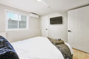 A bedroom with a bed, a window with blinds, an air conditioning unit, a television mounted on the wall, and a door.