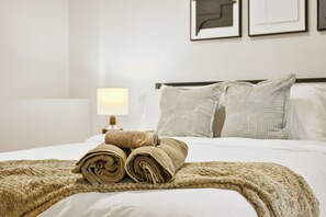 A neatly made bed with rolled towels and a blanket, a bedside lamp, and a wall with framed pictures. - Five75 Lux Prov PC Suite 3 (Providence)