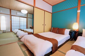 Ein traditionell eingerichtetes Hotelzimmer im japanischen Stil mit Tatami-Boden, einem großen Bett mit weißer Bettwäsche, einem kleinen Tisch mit Lampe und einer Schiebetür.