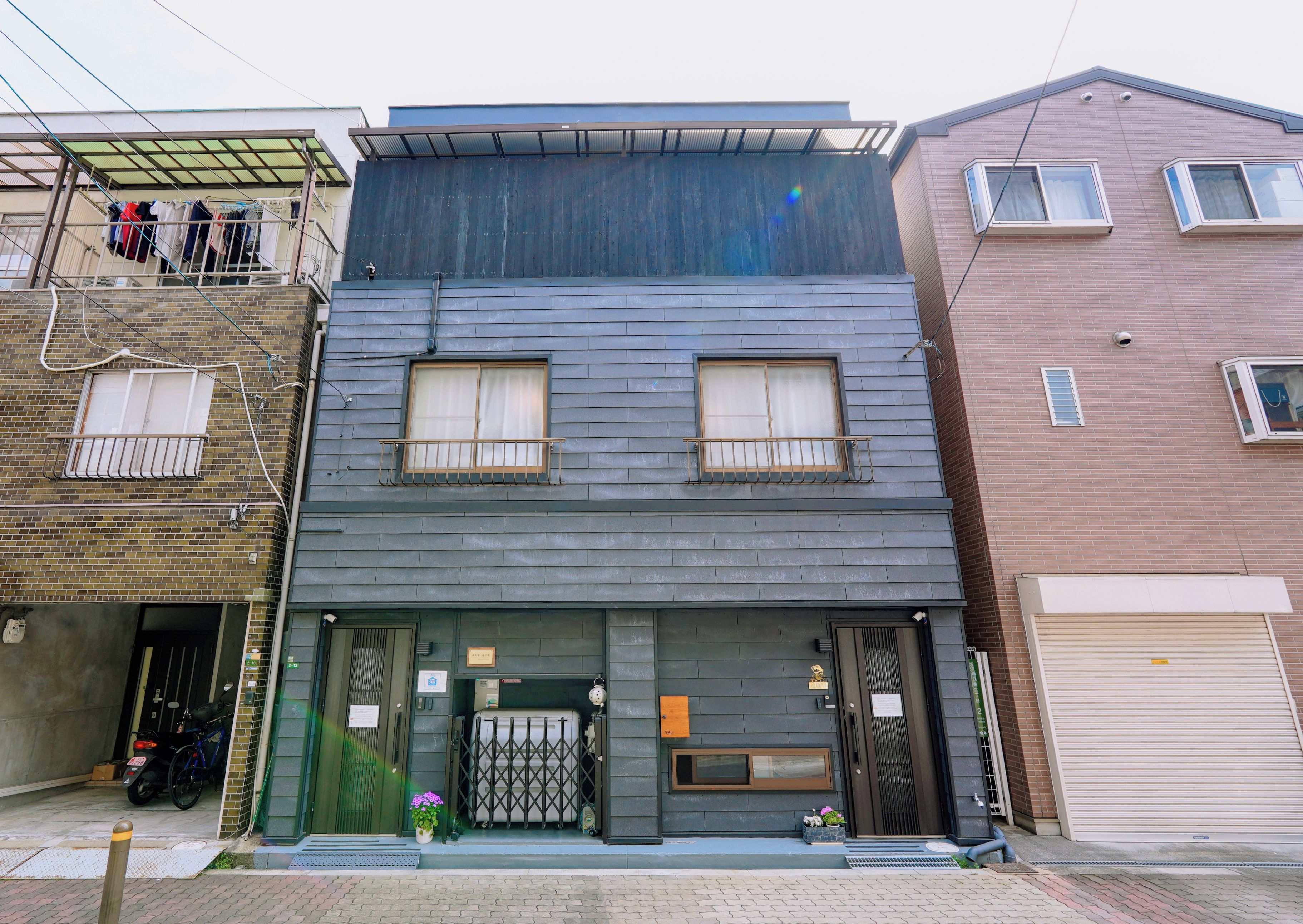 A two-story building with a dark facade, a gated entrance, and a small balcony with clothes drying.