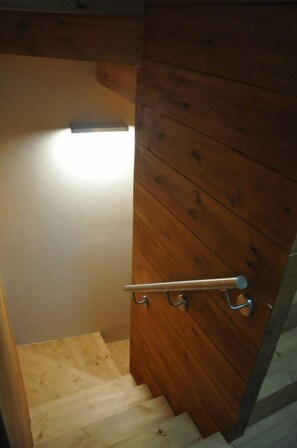 Un escalier en bois avec une rampe métallique, menant à une pièce où un luminaire est fixé au mur.