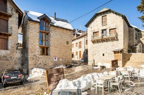 A stone building with a staircase leading to a second floor, outdoor seating area with tables and chairs, and a parked car. - El Pajar de la Leña - tucasaenlospirineos (Sandinies)