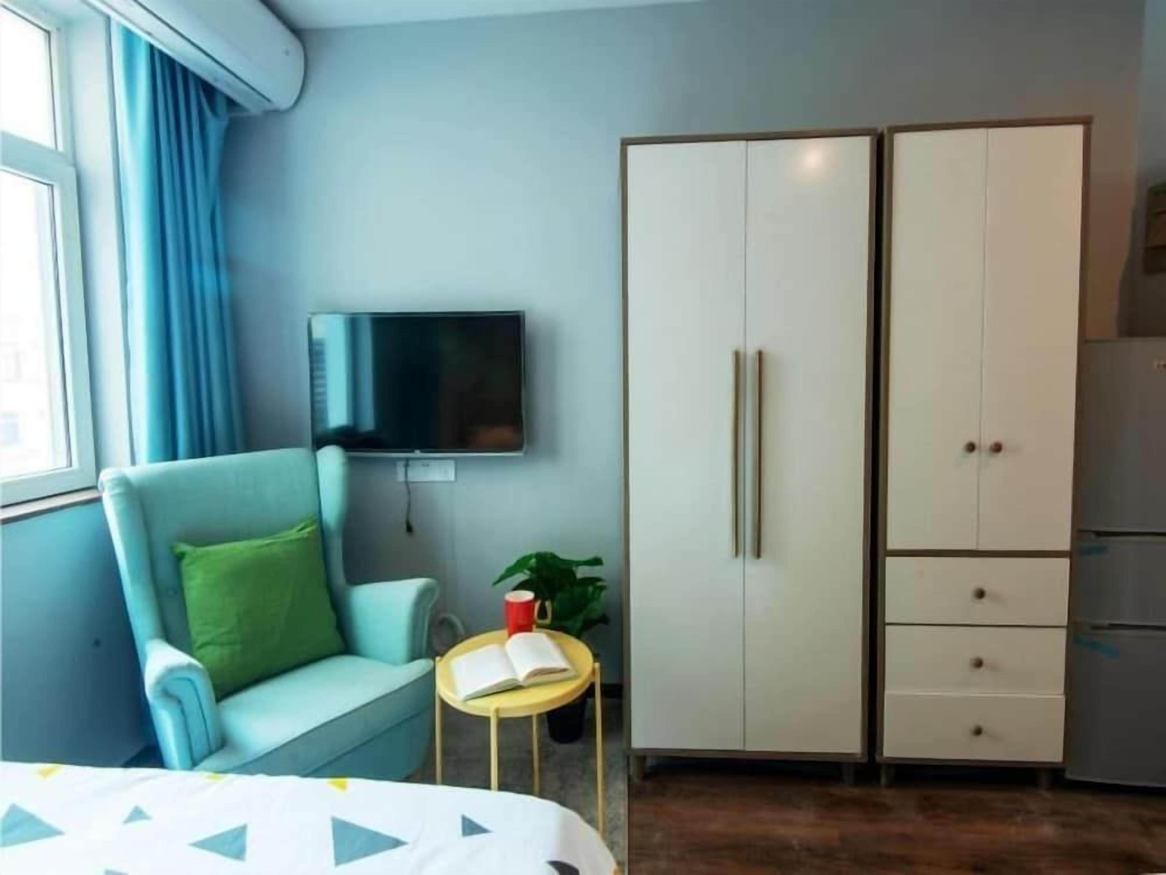 A room with a bed, a TV mounted on the wall, a wardrobe, a small table with a book, and a plant.