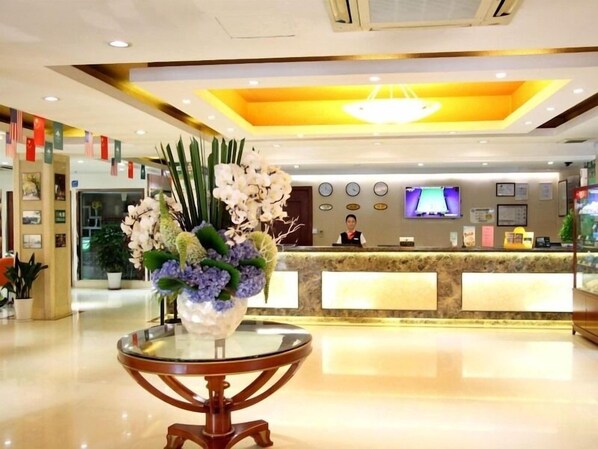 A hotel reception area with a glass-top table in the foreground, a reception desk with a monitor and a person behind it, and a chandelier above.